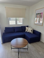 Un salón con un sofá seccional azul oscuro, una mesa de centro redonda de madera y un gran ventanal con persiana color beige.