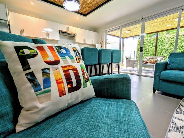A modern living room with teal sofas, a large window, and a view of greenery outside. - Upscale Tropical Retreat | 4BR in Gated Resort (Jaco)