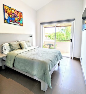 A bedroom with a bed, a nightstand, a painting, a balcony with a table and chairs, and a view of greenery outside. - Upscale Tropical Retreat | 4BR in Gated Resort (Jaco)