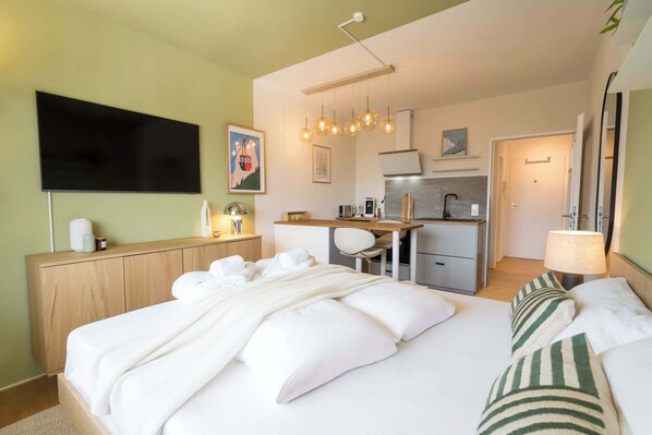 Habitación de hotel con cama, escritorio, silla, televisor y zona de cocina.