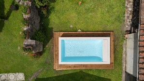 An aerial view of a rectangular swimming pool surrounded by a wooden deck, with a grassy area and some plants nearby.