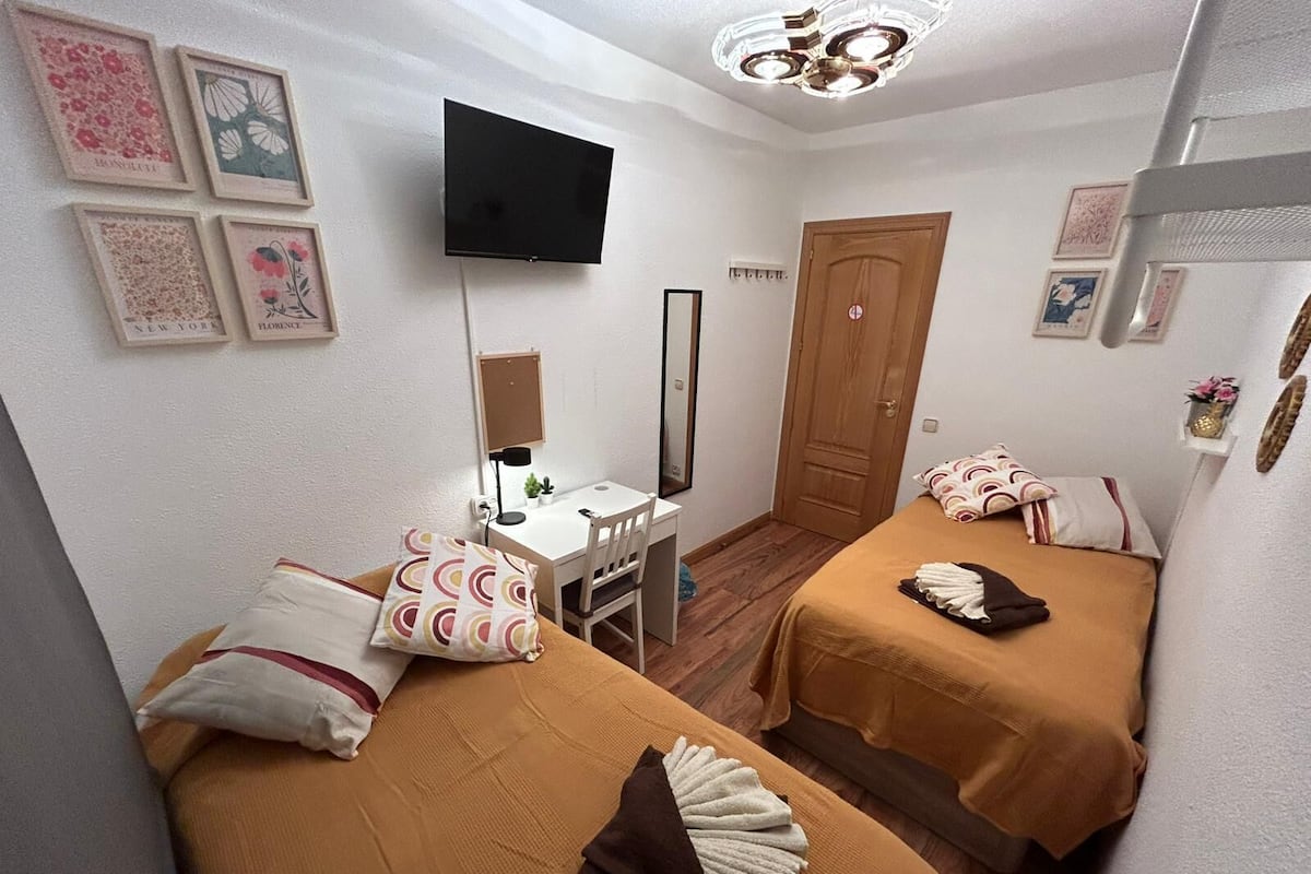 Habitación de hotel compacta con dos camas individuales, un escritorio con silla, un televisor de pantalla plana montado en la pared y una puerta de madera.