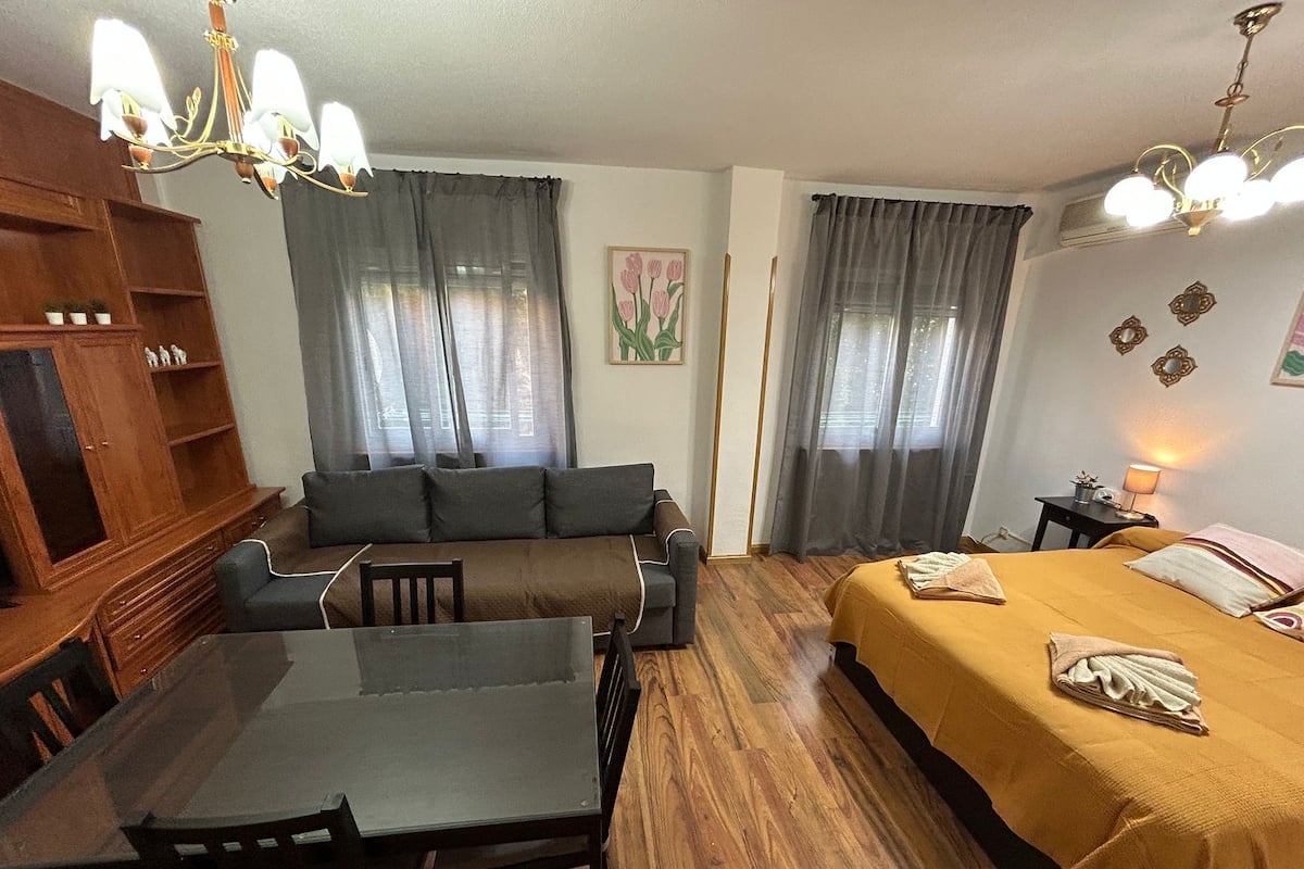 Una habitación acogedora con una mesa de comedor de madera, un sofá, una cama con colcha amarilla y una araña.