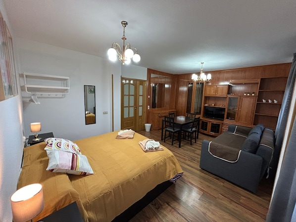 A room with a bed, a sofa, a dining table with chairs, a TV, wooden cabinets, and a chandelier. - Rooms Pontones (Madrid)