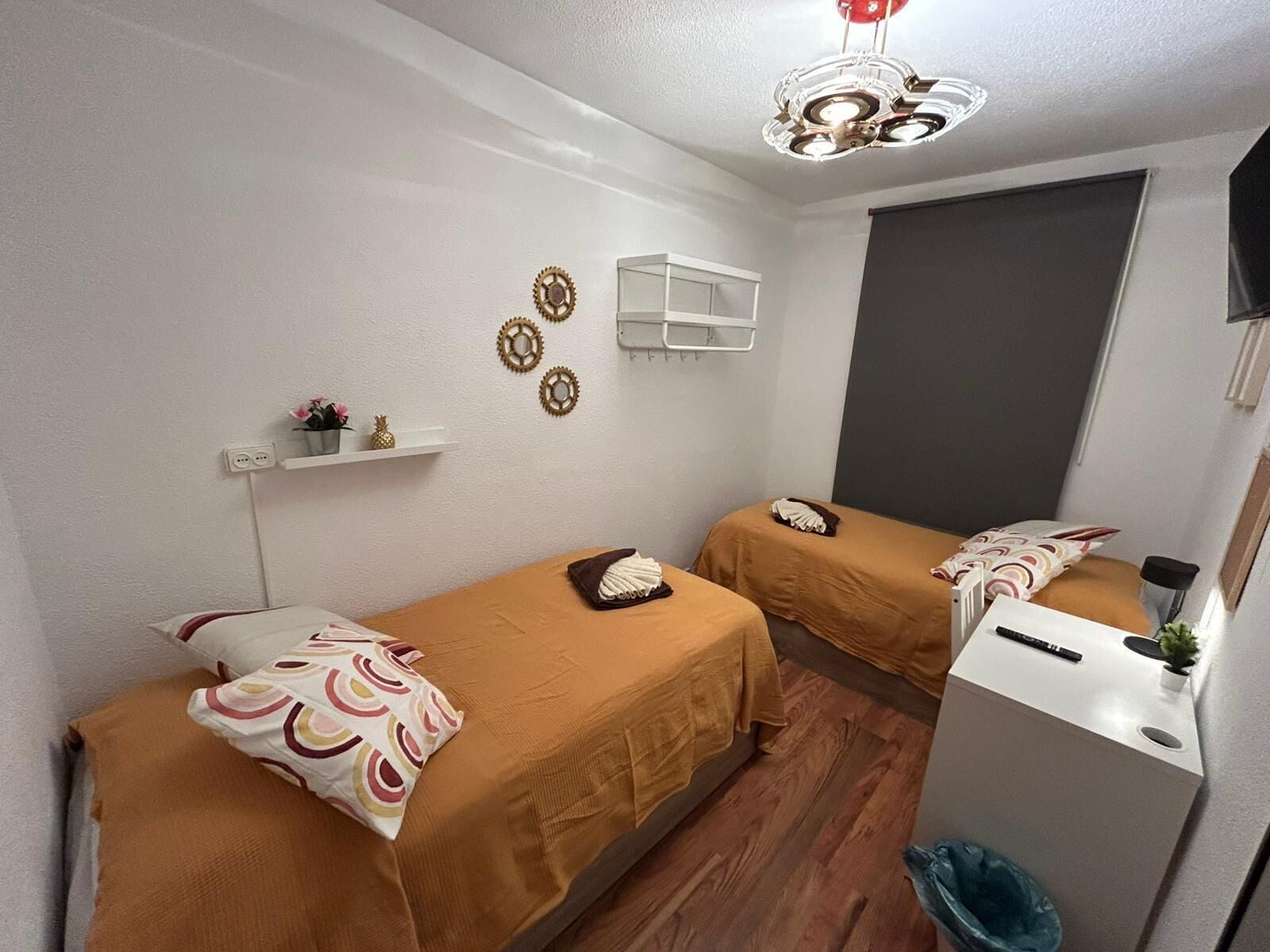 A small, clean room with two beds, a desk, a trash bin, and a TV on the wall.