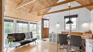 Een ruime woonruimte met een houten plafond, een zwarte bank, een eettafel met stoelen en grote ramen met uitzicht op het groen.