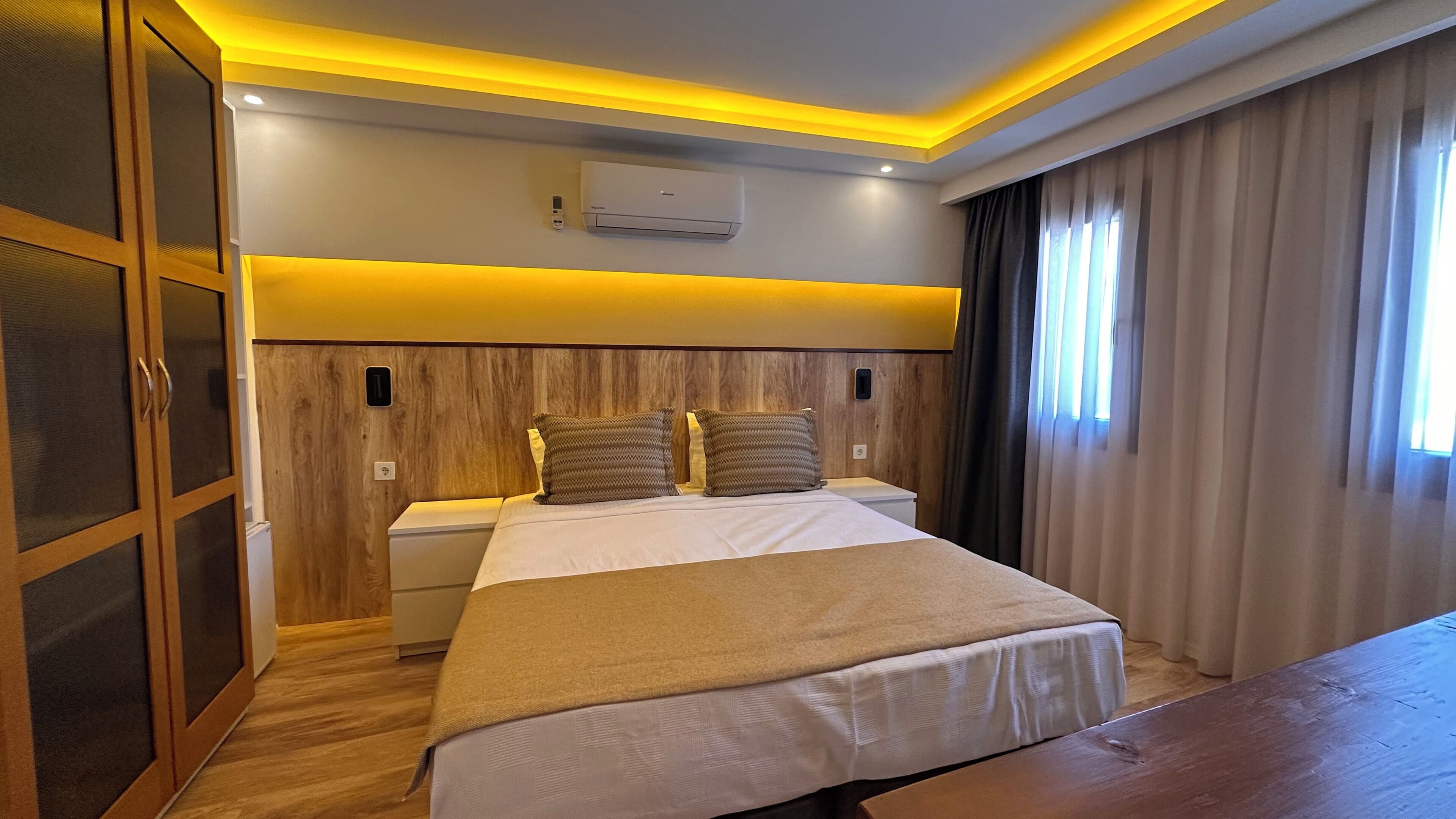 A hotel room with a wooden headboard, a bed with white linens, two pillows, a bedside table, and a window with curtains.