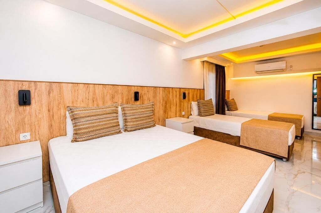 Ett modernt hotellrum med en stor säng, två mindre sängar, en sänggavel i trä, ett vitt nattduksbord, en nattlampa och en luftkonditioneringsanläggning.