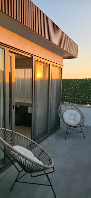 Un bâtiment moderne avec une entrée dotée d’une porte en verre, deux fauteuils en osier avec des coussins, et une vue dégagée sur le ciel.