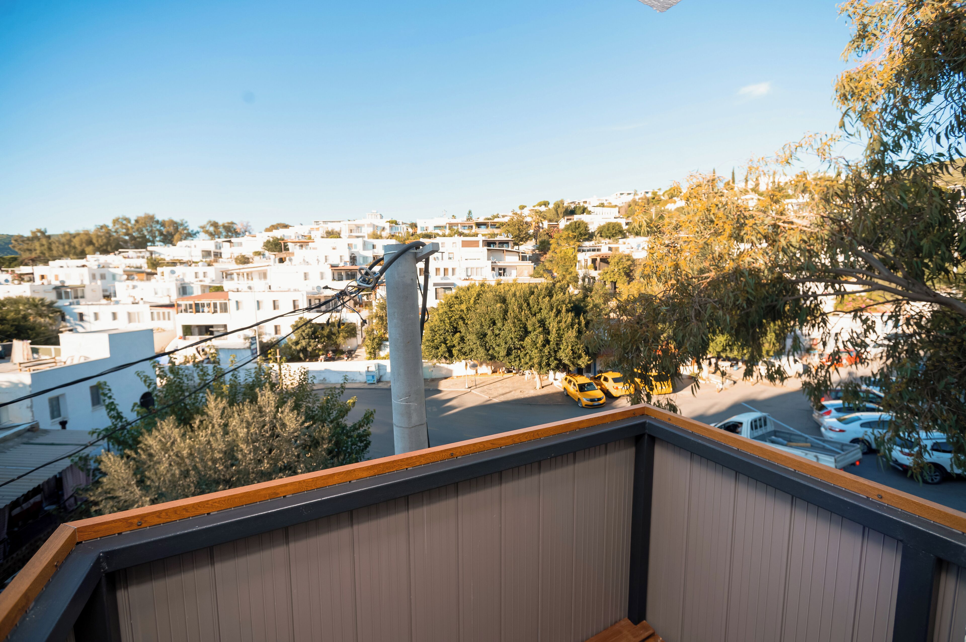 A view from a balcony overlooking a residential area with white buildings, a street with cars, and a tall utility pole.
