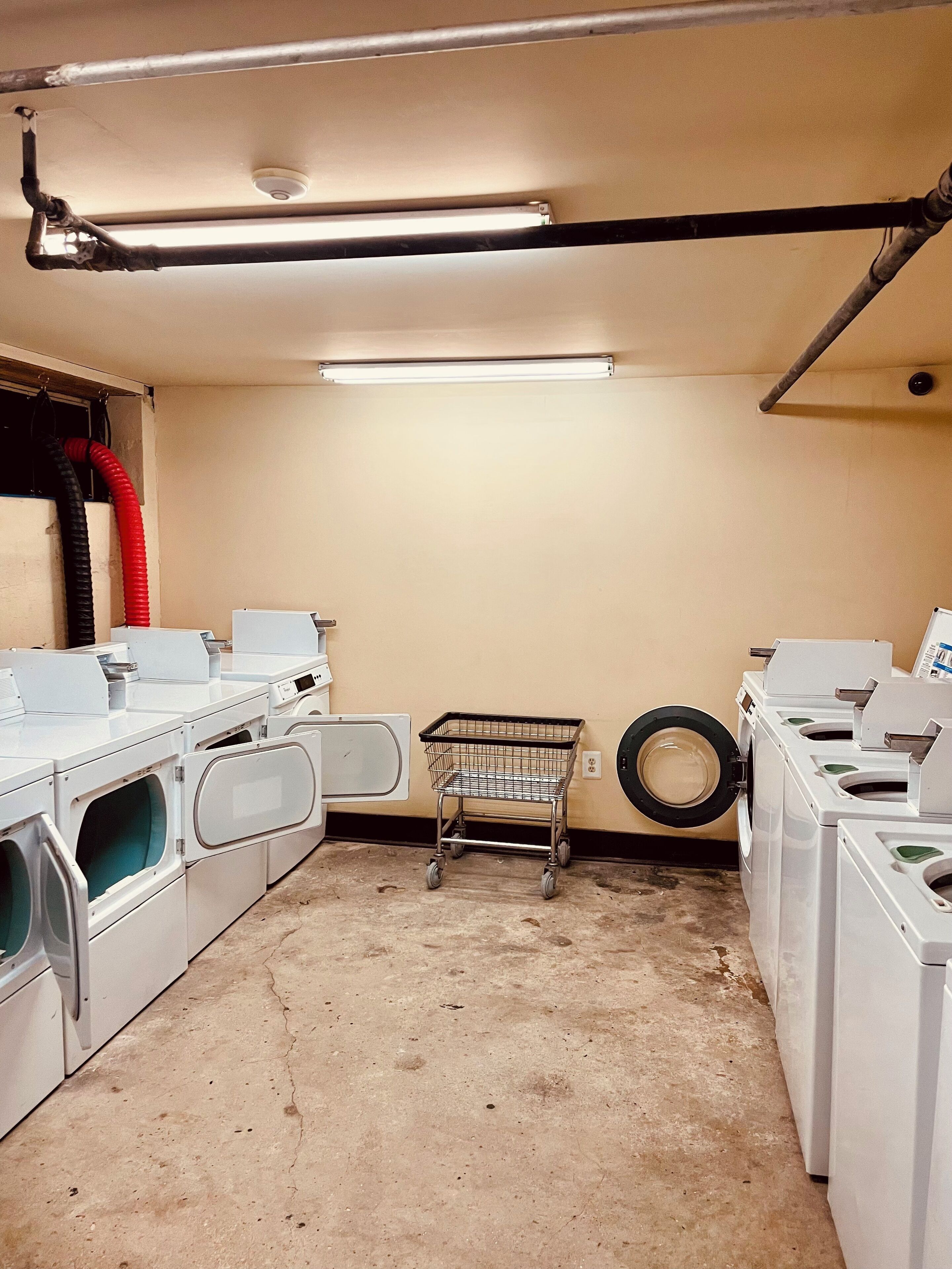 A laundry room with multiple washing machines, a metal laundry cart, and a tire against the wall.