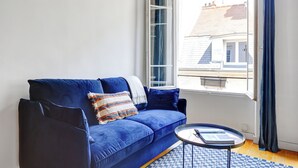 Un canapé bleu surmonté d’un plaid à motifs, une petite table ronde avec un livre dessus, et une fenêtre donnant sur des immeubles.