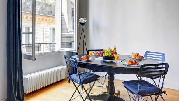 Un coin repas avec une table dressée pour deux personnes, agrémenté d’un panier de fruits, de croissants et d’un verre de jus d’orange.