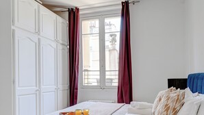 Une chambre avec un lit, une fenêtre avec des rideaux, un plancher en bois et un plateau de petit-déjeuner avec du jus d’orange.