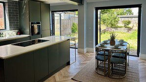 Uma cozinha moderna com uma ilha central, um espaço de refeições com uma mesa redonda e cadeiras, e vista para o quintal através de uma porta de vidro.