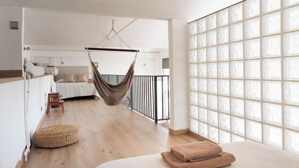Una habitación amplia con una silla colgante, una cama con toallas enrolladas, un taburete pequeño y un ventanal con diseño de cuadrícula.