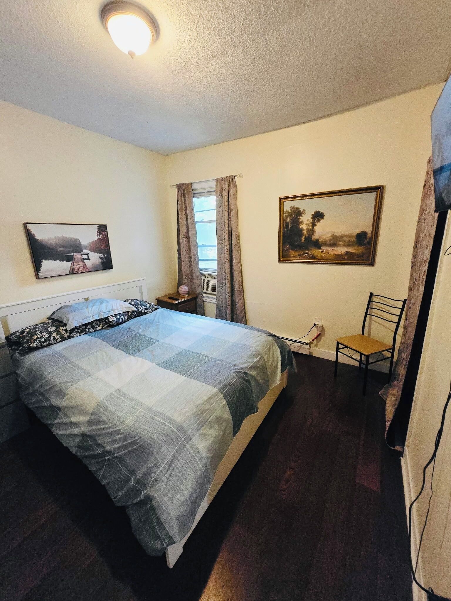 Une chambre à coucher avec un lit, un bureau, une chaise, une fenêtre avec des rideaux et deux tableaux accrochés au mur.