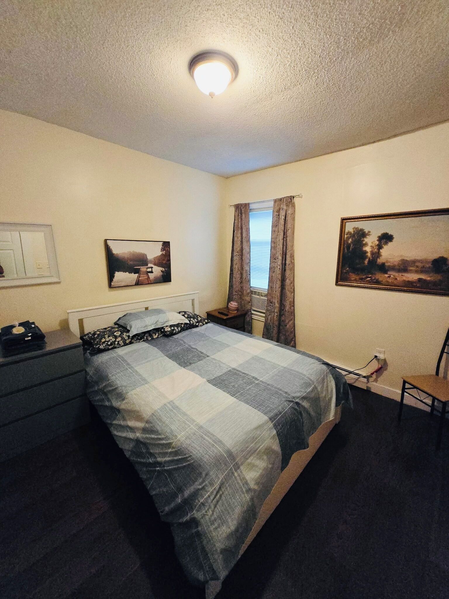 Een slaapkamer met een bed, nachtkastje, stoel, raam met gordijnen en twee aan de muur hangende schilderijen.