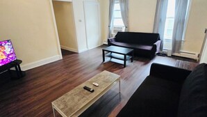 Una sala de estar con piso de madera oscura, un sofá negro, una mesa de centro y un televisor sobre un mueble.