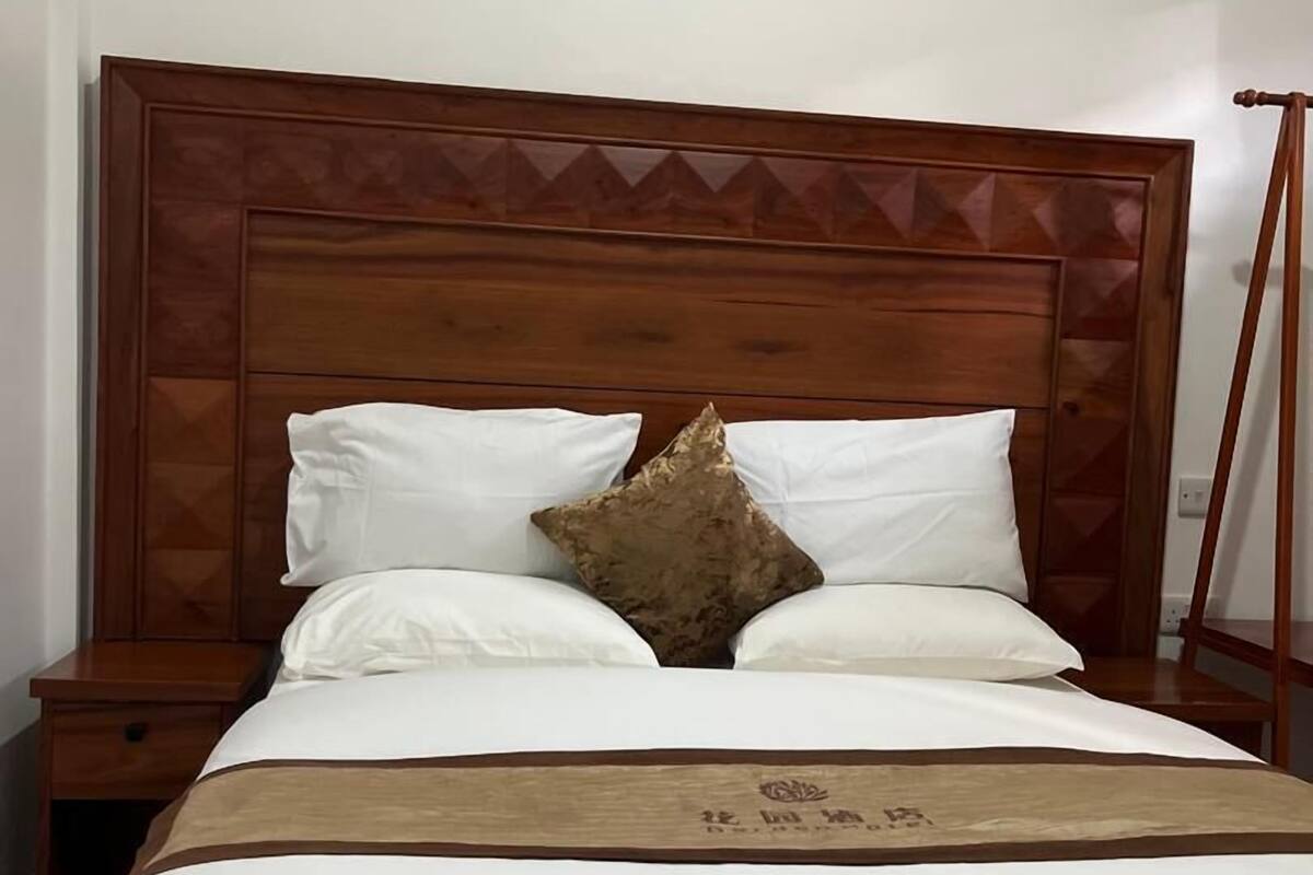 Un letto rifatto con lenzuola bianche e una coperta marrone, un poggiatesta in legno con motivo geometrico e un comodino in legno accanto.