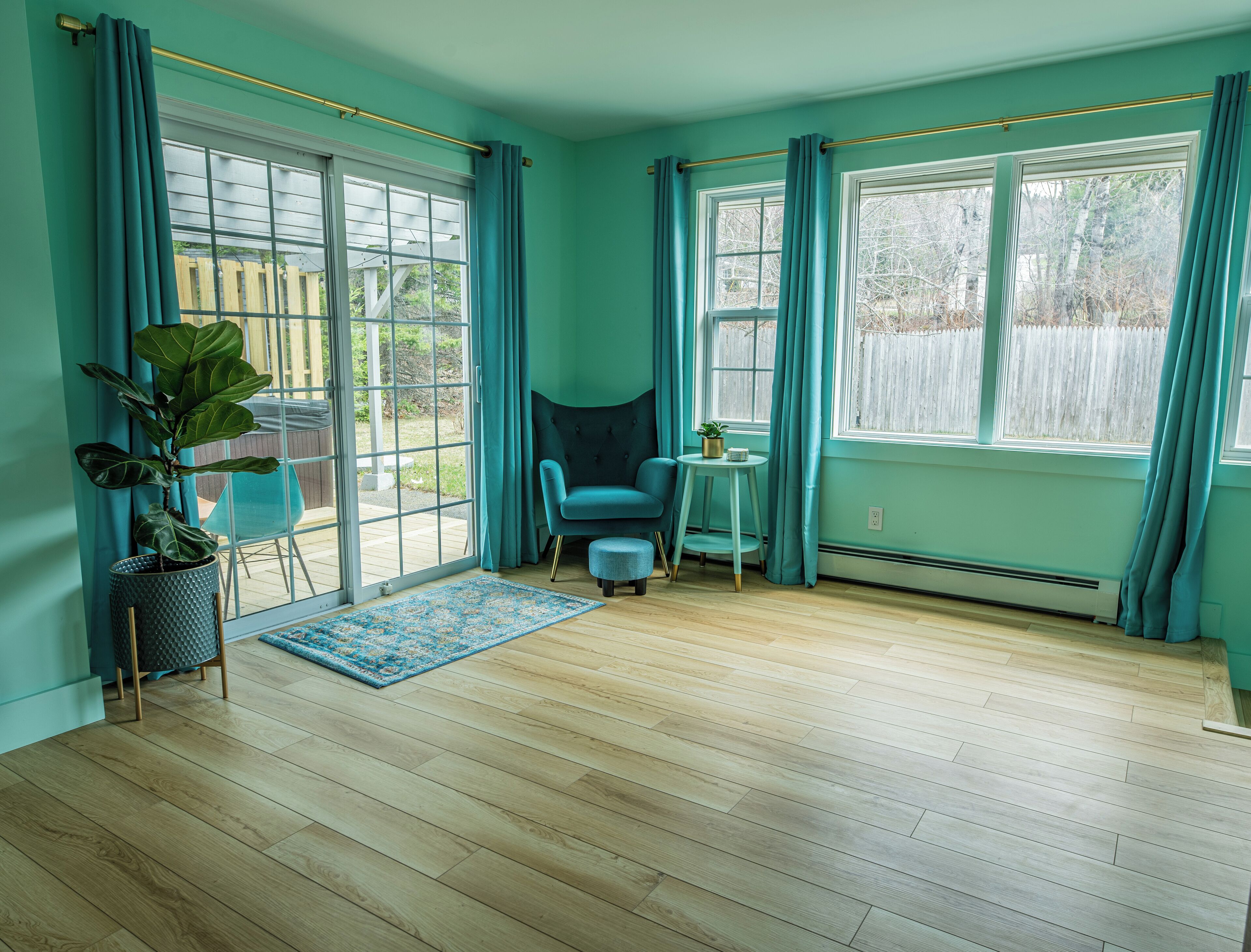 Una habitación con paredes verde azulado, piso de madera, una silla azul, una mesita con una planta y ventanas amplias con cortinas verde azulado.