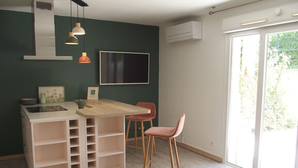 Une cuisine moderne avec un îlot central en bois, un coin repas agrémenté d’une chaise rose et une grande fenêtre donnant sur un paysage verdoyant.