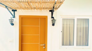 Eine gelbe Tür mit Gittermuster, begleitet von zwei schwarzen Wandleuchtern, und ein Fenster mit horizontalen Rollläden zur Rechten.