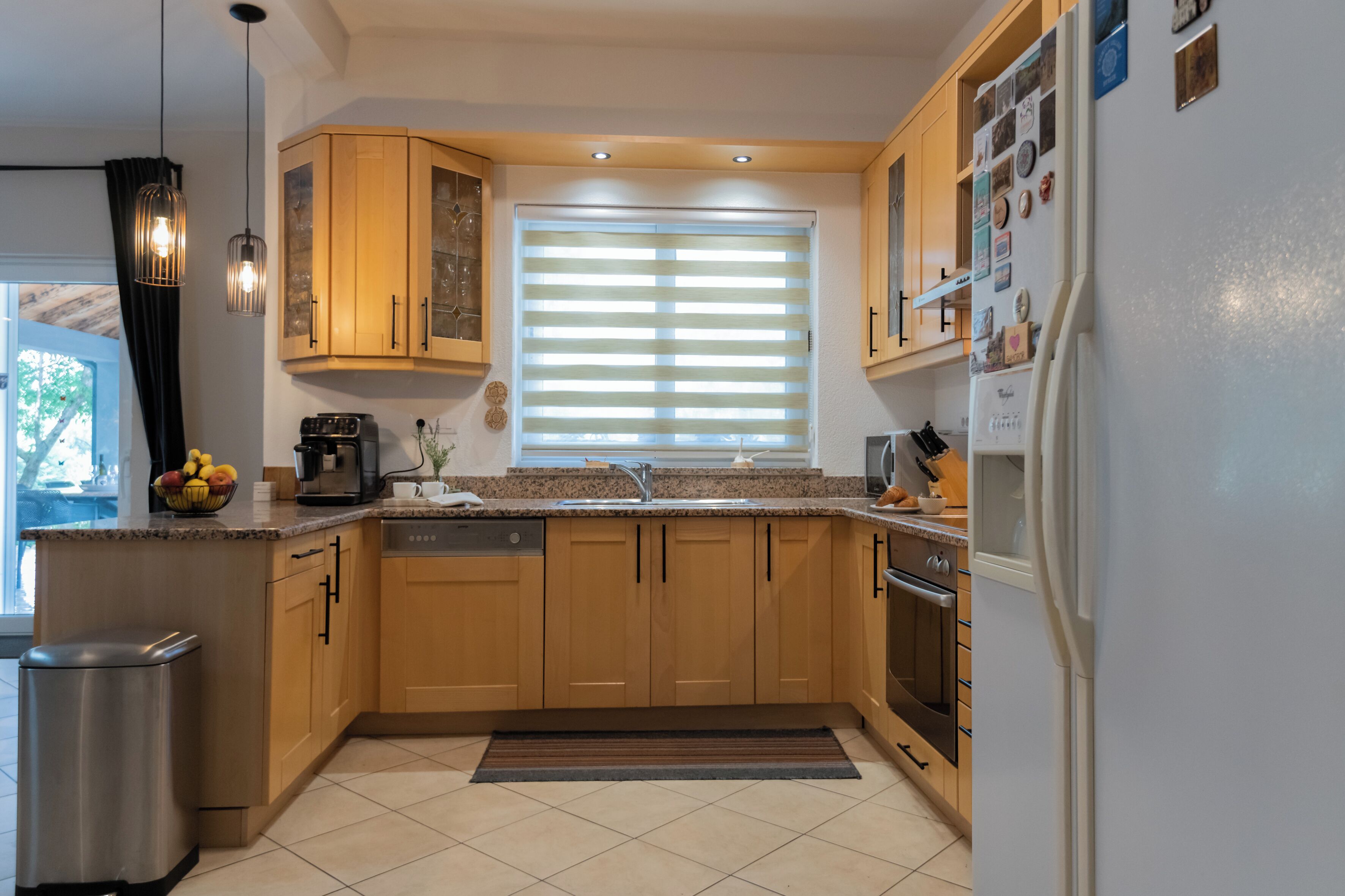 A modern kitchen with wooden cabinets, a built-in dishwasher, a refrigerator, and a window with blinds.