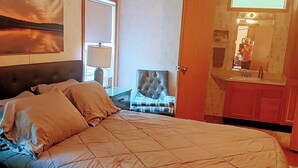 Ein Schlafzimmer mit einem großen Bett, einem Sessel, einem Holzkopfende, einem Deckenventilator und einem durchgehenden Türspalt sichtbaren Badezimmer.