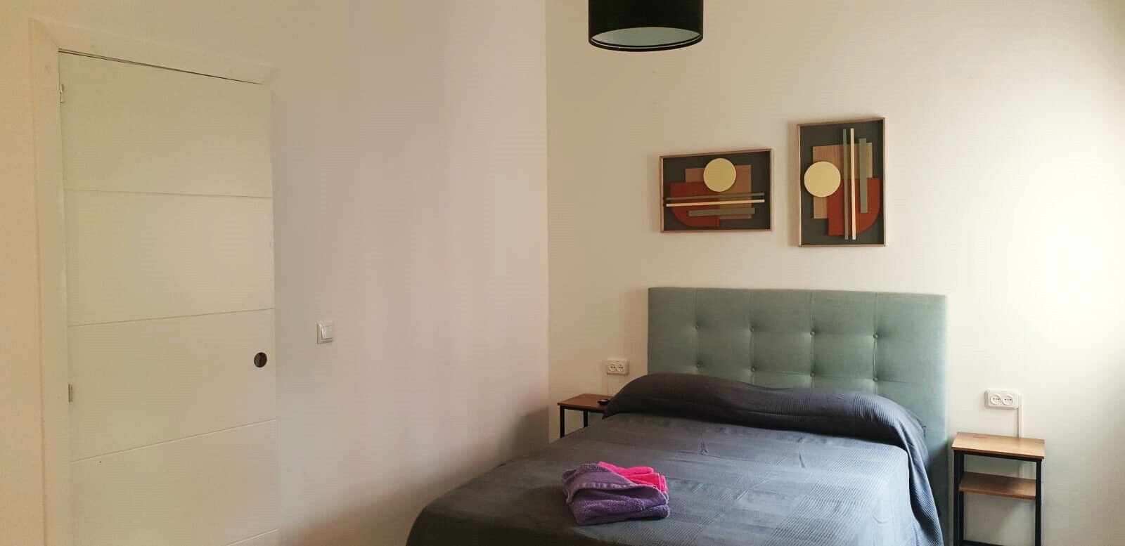Un dormitorio con un cabecero tapizado de color verde, una cama con cubrecama oscuro, dos mesitas de noche y un cuadro abstracto en la pared.