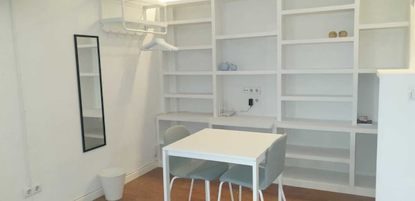 Una habitación con paredes blancas, una mesa blanca y sillas a juego. Se observa una estantería blanca con algunos objetos encima.