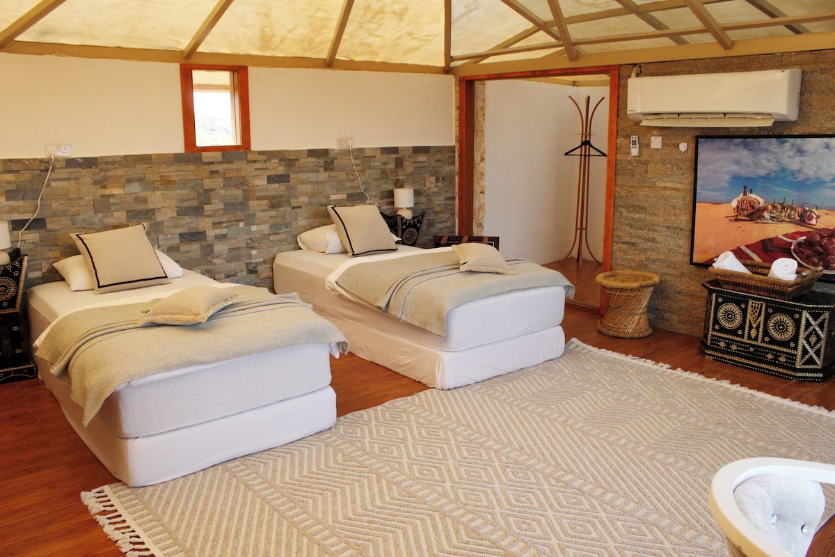 A room with two beds, a patterned rug, a small round table, and a wall-mounted air conditioner.