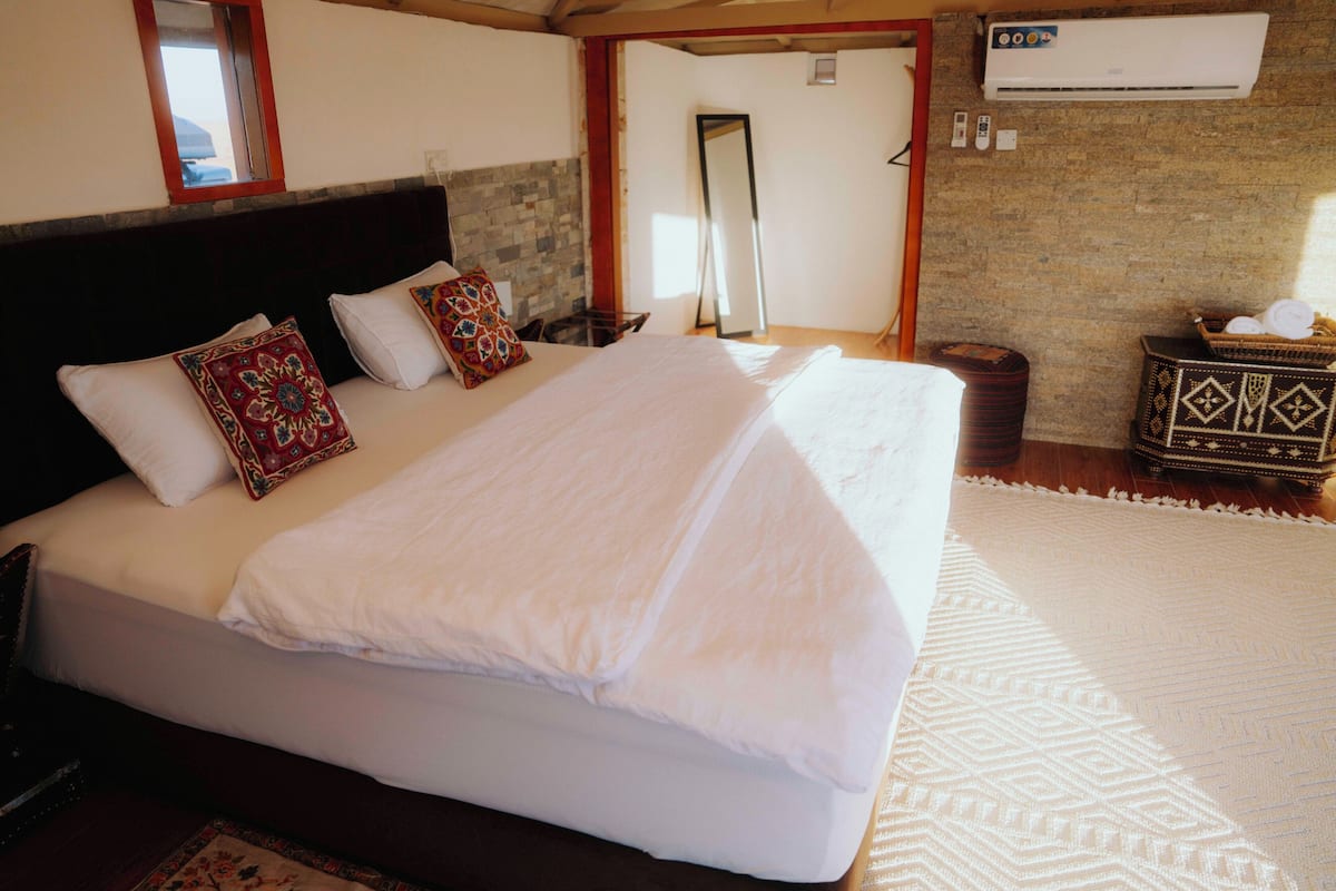A neatly made bed with white linens and decorative pillows, a wooden headboard, a woven rug, and a small side table with a basket.