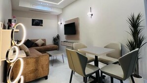 A modern living room with a sofa, a dining table with chairs, a TV mounted on the wall, and a potted plant. - Princess Plaza hotel (Ma'an)