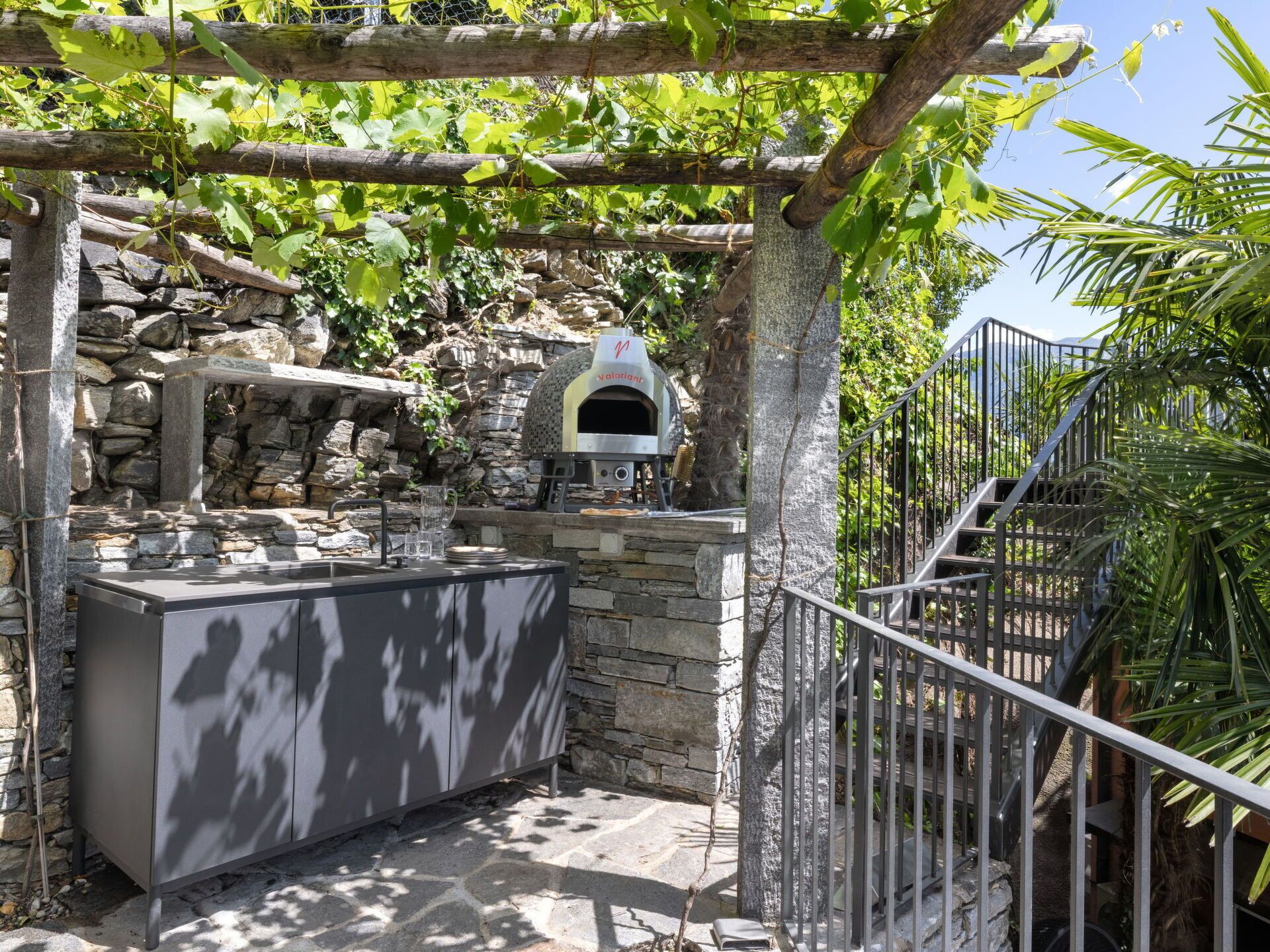 An outdoor kitchen area with a stone wall, a pizza oven, and a metal counter.