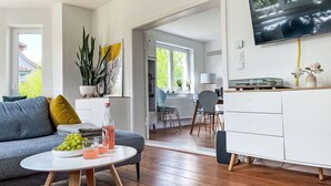 Un salon moderne avec un canapé gris, une table basse ronde sur laquelle sont disposés des fruits et des boissons, un meuble blanc et un téléviseur fixé au mur.