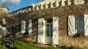 A stone building with white shutters, a small porch with chairs, and a stone tower. - 
Ancien Haras de la Tour Chambre Le Parc (Lamothe-Landerron)