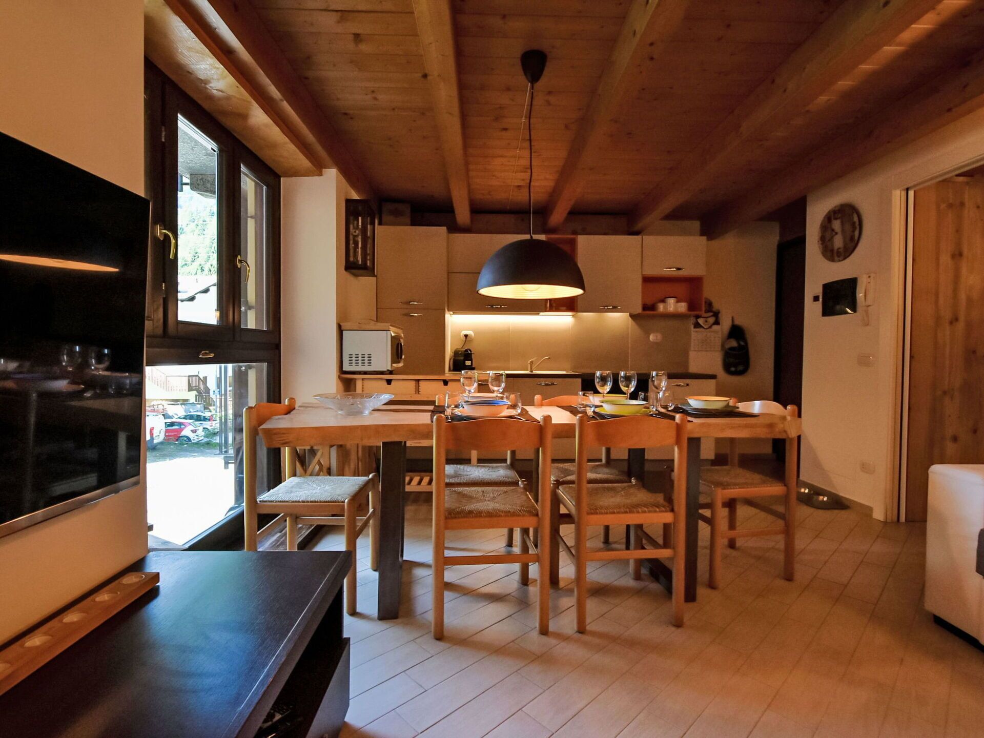 Une cuisine moderne avec des poutres en bois, une table à manger dressée pour six personnes et une grande fenêtre donnant sur un parking.