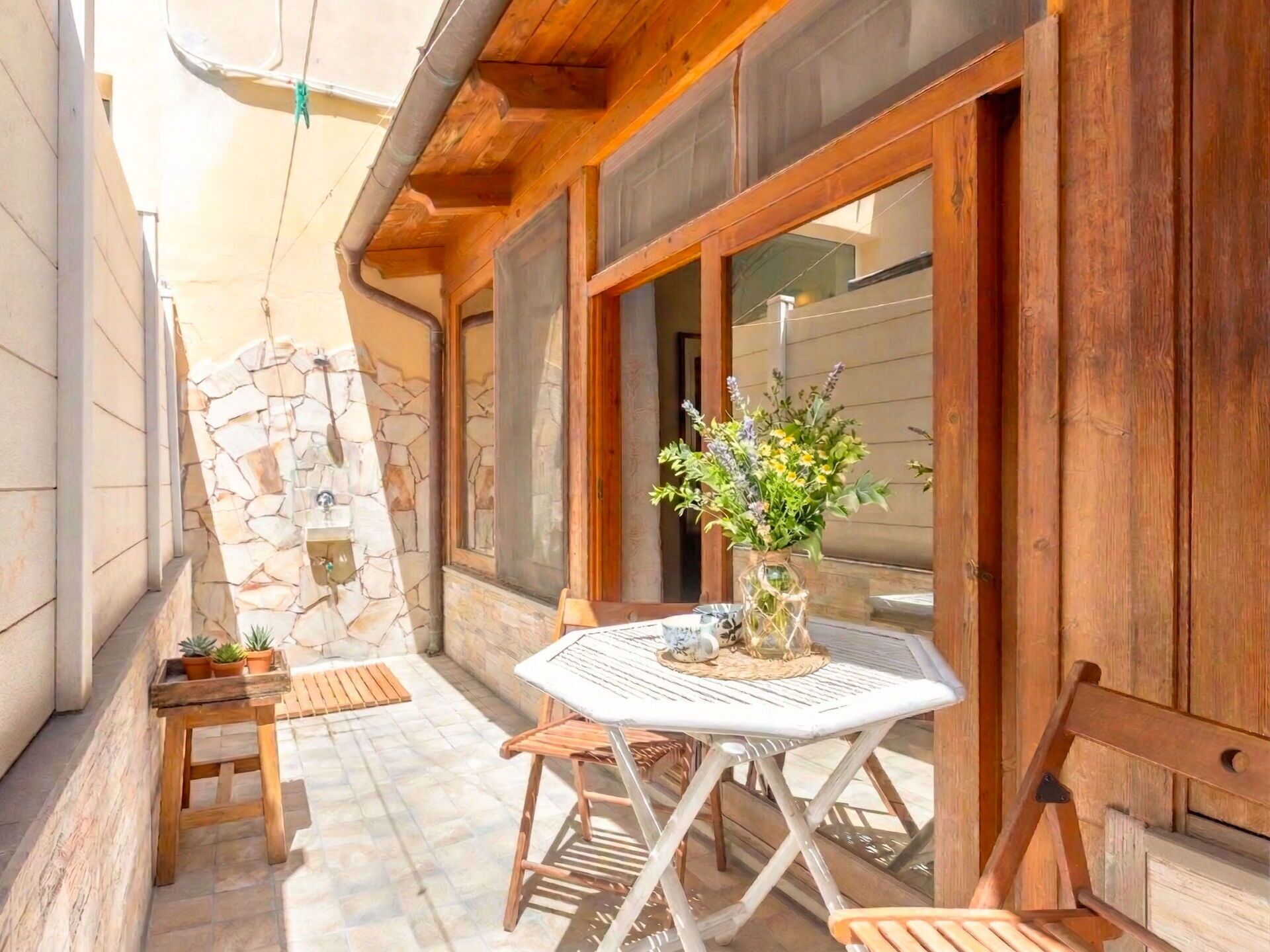 A wooden exterior with a stone wall, a table with chairs, and a vase of flowers.