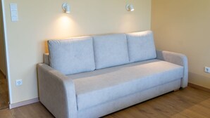A light grey sofa with two cushions against a plain wall, two wall-mounted lights, and a wooden floor. - Beautiful apartment for 3 guests with A/C, WIFI, TV and balcony (Strobl)