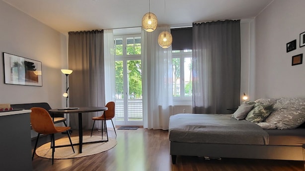 A modern bedroom with a bed, a desk, and a chair. There are large windows with curtains, a floor lamp, and a ceiling light. - Nice apartment for 2 guests with TV and balcony (Magdeburg)