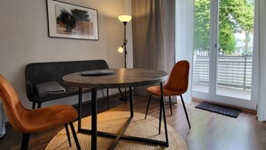 A modern living room with a round wooden table, two orange chairs, a black sofa, a floor lamp, and a large window with a view of trees and buildings. - Nice apartment for 2 guests with TV and balcony (Magdeburg)