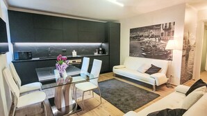A modern living room with a glass dining table, white sofas, and a kitchen area with dark cabinetry and a marble backsplash.