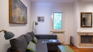 Ett vardagsrum med en grå soffa, en tavla, ett litet inramat foto, ett fönster med persienner och en öppen spis.