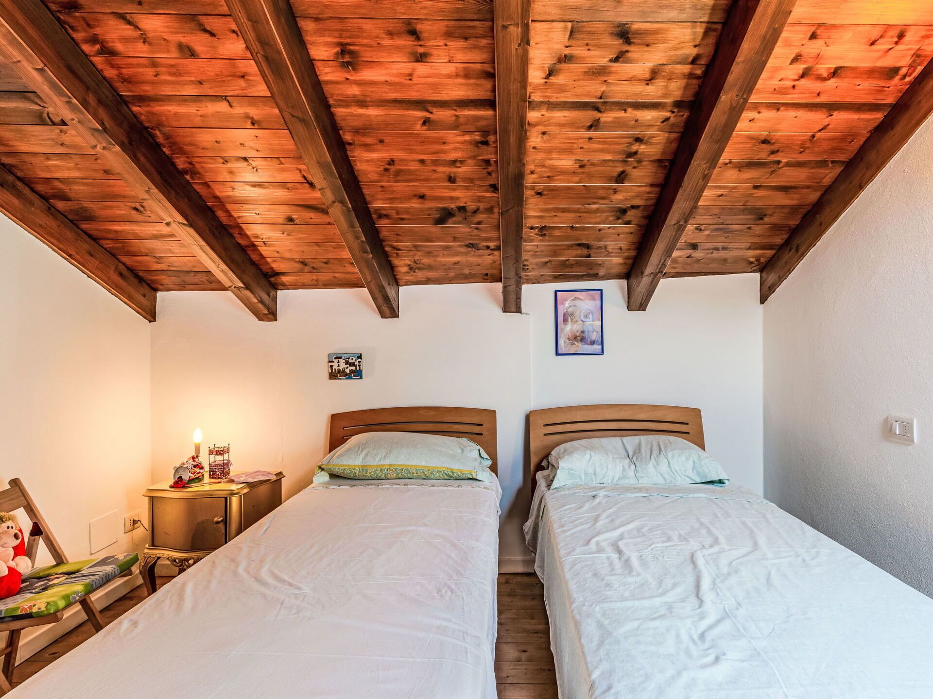 A bedroom with two beds, wooden ceiling beams, a small bedside table with a lamp, and a framed picture on the wall.