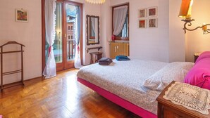 A bedroom with a bed, a wooden floor, a chandelier, a radiator, a window with curtains, and a wall-mounted lamp.