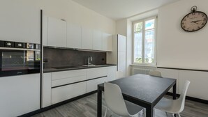 Una cocina moderna con armarios blancos, encimera negra y horno integrado. Hay una mesa de comedor con dos sillas blancas y un reloj en la pared.