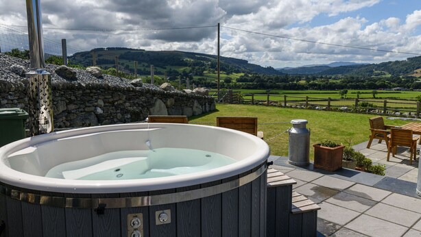 A hot tub with a stone wall backdrop, surrounded by a patio with chairs and a table.