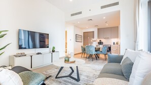 A modern living room with a sofa, a coffee table, and a TV. There is a dining area with chairs and a kitchen in the background. - Creek Views 1BR, Short Drive to Ras Al Khor Sanctuary and Festival City Mall (Dubai)