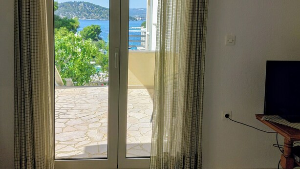 A room with a sliding glass door leading to a balcony with a stone floor and a view of a body of water and greenery. - Apartment A2 dvije spavaće sobe, Garden Terrace 40 m2 with Outdoor Shower (Ražanj)